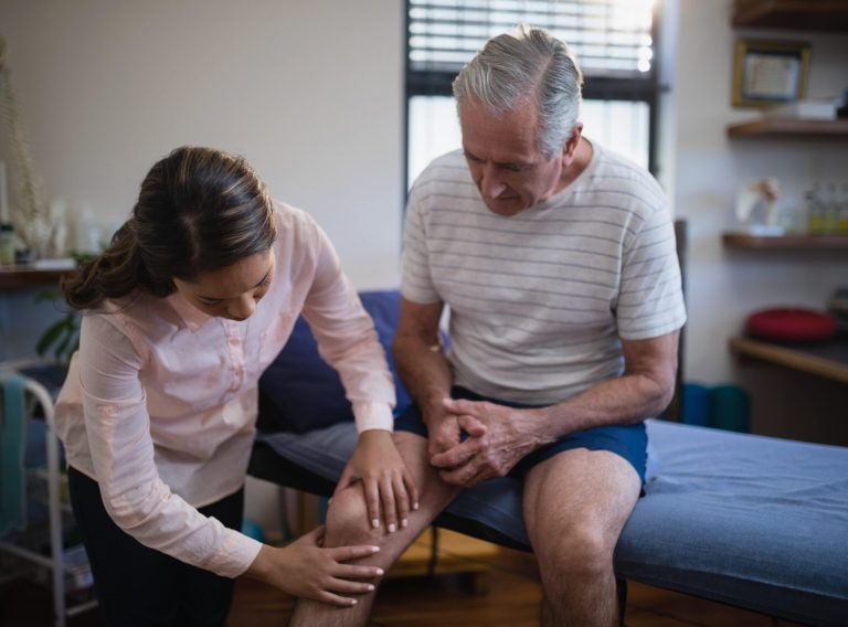 female-therapist-examining-knee-of-senior-male-pat-7FQHTEU-min